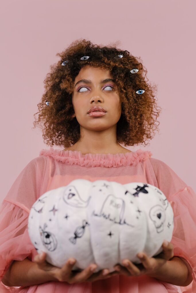 Woman with scleral contact lenses which cover her eyes so they appear entirely white. She is holding a white pumpkin decorated with ghosts and pumpkins.